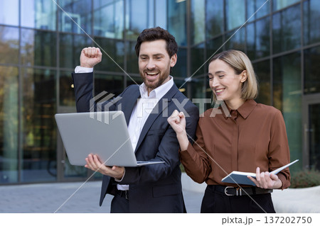 Happy business colleagues celebrating an achievement, standing with a laptop and documents outside an modern office building, representing teamwork and successful partnership Happy business colleagues celebrating an achievement, standing with a laptop and documents outside an modern office building, representing teamwork and successful partnership 137202750