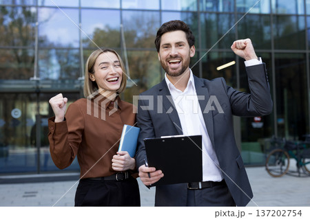 Two excited business colleagues celebrating a professional victory, raising their fists in triumph while standing outside a modern office building, representing teamwork and accomplishment 137202754