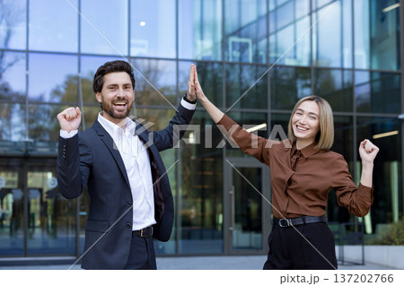 Two cheerful business people, a man and a woman, standing outside an office building, celebrating achievement and a successful partnership with a high five gesture 137202766