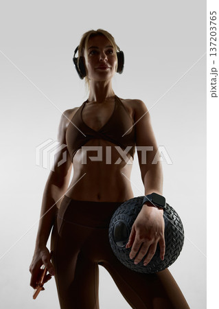 Woman holding textured fitness ball wearing smartwatch and headphones in studio. Woman holding textured fitness ball wearing smartwatch and headphones in studio. 137203765