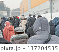 People standing in queue during snowfall near urban building, wearing winter jackets and hoods, backs turned to camera in cold weather. Concept of queue, waiting line, social issue, crowd 137204535