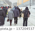 People waiting at transport stop during snowfall in city, wearing winter jackets on snow covered street near building. Concept of transport stop, public transport, commuting, urban mobility 137204537