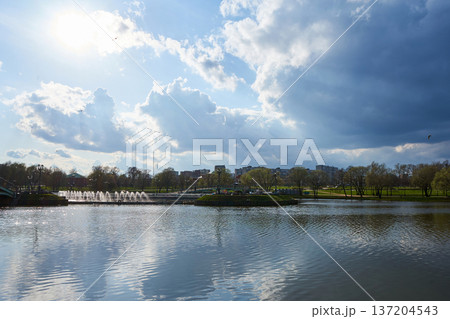 Scenic view of a large pond with fountains in a city park under a partly cloudy sky, showing a calm water surface reflecting the sky and surrounding trees. Scenic view of a large pond with fountains in a city park under a partly cloudy sky, showing a calm water surface reflecting the sky and surrounding trees. 137204543