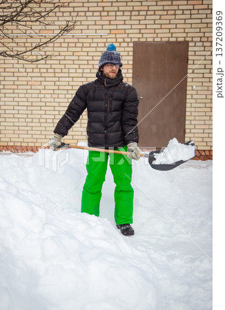 Man in winter jacket shoveling snow with shovel in front of brick house after heavy snowfall 137209369