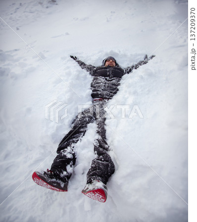 Adult man lying on fresh snow making snow angel with outstretched arms during winter day outdoors 137209370