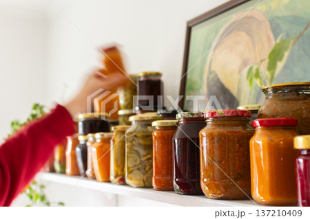 A person holds a jar of canned vegetables while checking the shelf filled with various jars of preserved food. This activity takes place during winter preparations for storage A person holds a jar of canned vegetables while checking the shelf filled with various jars of preserved food. This activity takes place during winter preparations for storage 137210409