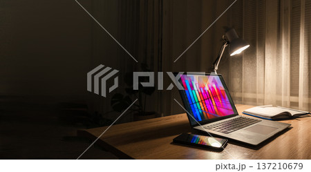 A desk lamp illuminates a laptop on a wooden table at night, displaying a colorful histogram and data graph A desk lamp illuminates a laptop on a wooden table at night, displaying a colorful histogram and data graph 137210679