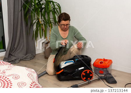 In a living room, a person is getting a vacuum cleaner ready for cleaning. The person is seated on the floor with the vacuum cleaner open and looks focused on the task In a living room, a person is getting a vacuum cleaner ready for cleaning. The person is seated on the floor with the vacuum cleaner open and looks focused on the task 137213144