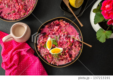 A vibrant bowl of cold beetroot soup features hard-boiled eggs and fresh herbs. A lemon slice accompanies it, all set against a black surface with a pink cloth for contrast A vibrant bowl of cold beetroot soup features hard-boiled eggs and fresh herbs. A lemon slice accompanies it, all set against a black surface with a pink cloth for contrast 137213191