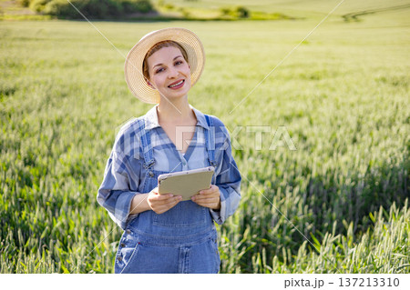 A young woman wearing overalls and a straw hat stands in a green field holding a tablet computer and smiling A young woman wearing overalls and a straw hat stands in a green field holding a tablet computer and smiling 137213310