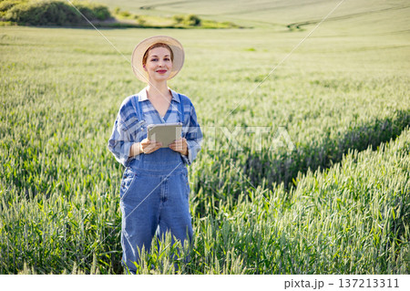 A woman in overalls and a straw hat stands in a green field holding a tablet computer and smiling A woman in overalls and a straw hat stands in a green field holding a tablet computer and smiling 137213311