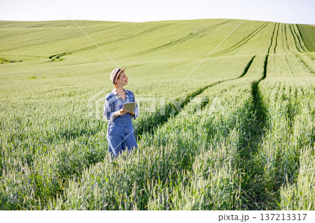 A woman in overalls and a hat stands in a lush green wheat field holding a tablet and looking thoughtfully into the distance A woman in overalls and a hat stands in a lush green wheat field holding a tablet and looking thoughtfully into the distance 137213317