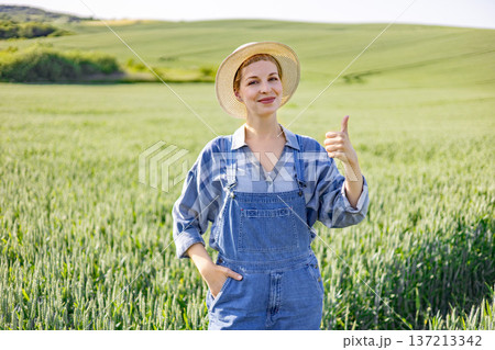 A smiling woman wearing overalls and a straw hat gives a thumbs-up gesture while standing in a lush green field of crops under a clear sky A smiling woman wearing overalls and a straw hat gives a thumbs-up gesture while standing in a lush green field of crops under a clear sky 137213342