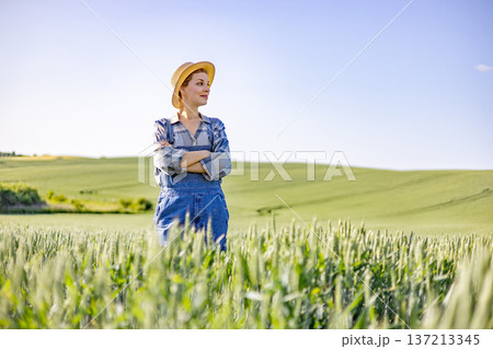 A confident woman wearing overalls and a straw hat stands with her arms crossed in a lush green wheat field under a clear blue sky 137213345
