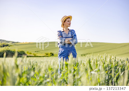 A confident woman wearing overalls and a straw hat stands with her arms crossed in a vast green wheat field under a clear blue sky 137213347