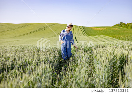 A young woman wearing overalls and a hat walks through a vast green wheat field under a clear blue sky 137213351