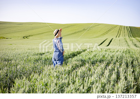 A young woman wearing denim overalls and a straw hat stands in a vast green wheat field under a clear blue sky, shielding her eyes from the sun A young woman wearing denim overalls and a straw hat stands in a vast green wheat field under a clear blue sky, shielding her eyes from the sun 137213357