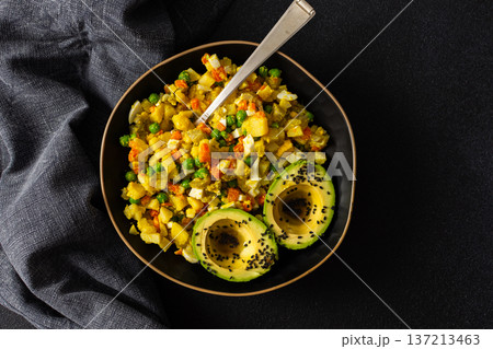 A colorful salad combines boiled egg, tender chicken breast, diced potatoes, vibrant carrots, and fresh green peas, topped with creamy avocado and sesame seeds, all drizzled with olive oil 137213463