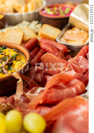 A vibrant charcuterie board showcases a variety of cheese, sausages, hummus, nuts, and sandwiches. Friends gather at a sunny outdoor picnic, enjoying delicious food together 137213511