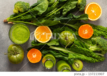 Fresh spinach, chard, and broccoli sit among sliced oranges and tangerines as juice is prepared in a kitchen. The scene captures a vibrant approach to healthy drinking 137213907