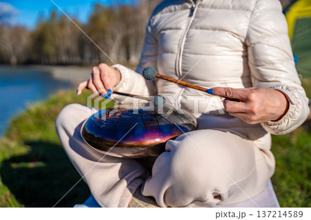 Close-up of hands playing a steel tongue drum with mallets against the backdrop of Altai mountains and river. Concept of sacred sound, touch of nature, and mindful musical ritual Close-up of hands playing a steel tongue drum with mallets against the backdrop of Altai mountains and river. Concept of sacred sound, touch of nature, and mindful musical ritual 137214589