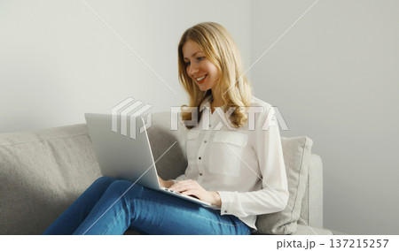 Happy modern woman working with laptop, freelancer girl sitting on a couch in the living room Happy modern woman working with laptop, freelancer girl sitting on a couch in the living room 137215257