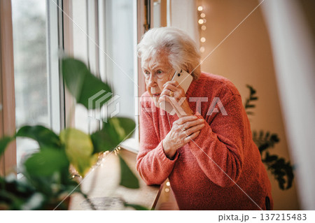 Old woman standing by window, making phone call. 137215483