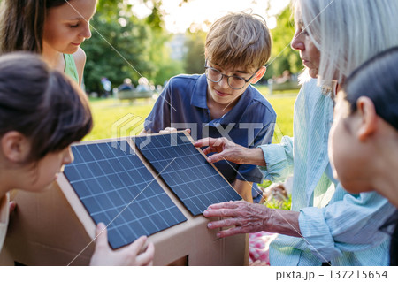 Children learning about renewable energy and solar panels during sustainable education class outdoors, using cardboard model of house wit solar panel on roof. Children learning about renewable energy and solar panels during sustainable education class outdoors, using cardboard model of house wit solar panel on roof. 137215654
