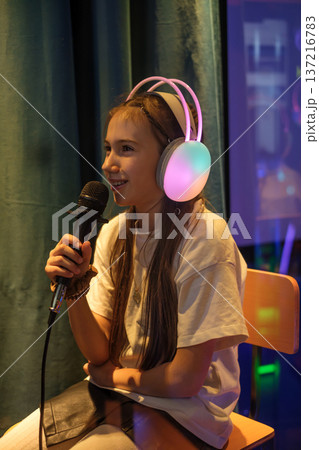 A girl in luminous headphones sings into a microphone at a karaoke club. The child gives an interview in a TV and radio company 137216783