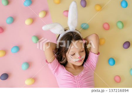 A cute, beautiful girl with makeup in a rabbit costume lies on a pastel pink background with Easter eggs 137216894
