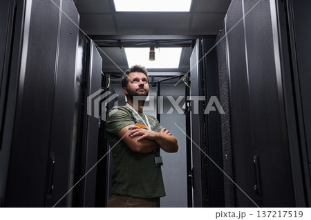 Data center technician standing between server racks with arms crossed looking up while monitoring equipment 137217519