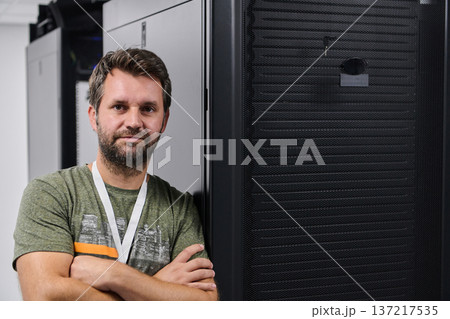 IT technician standing in front of server rack in a data center with arms crossed and confident smile IT technician standing in front of server rack in a data center with arms crossed and confident smile 137217535