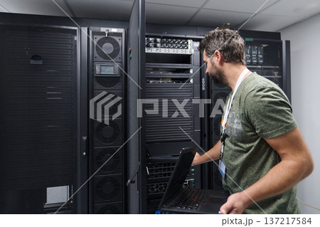 IT technician working on server rack in data center with laptop performing network maintenance 137217584