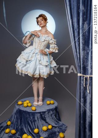 Young woman in vintage dress standing on pedestal with lemons in studio with blue curtain Young woman in vintage dress standing on pedestal with lemons in studio with blue curtain 137218164