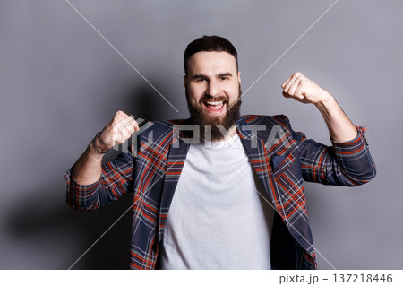 Excited bearded man very glad to success. Young cheerful guy in checkered jacket smiling at camera and gesturing with hands, gray studio background Excited bearded man very glad to success. Young cheerful guy in checkered jacket smiling at camera and gesturing with hands, gray studio background 137218446