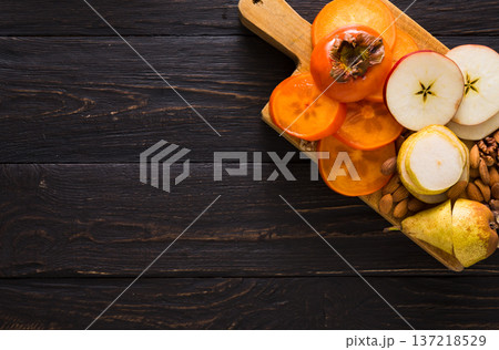 Healthy food - wood board with cut organic raw fruits on wooden background. Top view of sliced ingredients for fruit salad or snack - apple, pear, nuts, persimmon. Flat lay, copy space Healthy food - wood board with cut organic raw fruits on wooden background. Top view of sliced ingredients for fruit salad or snack - apple, pear, nuts, persimmon. Flat lay, copy space 137218529