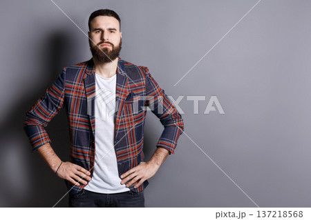 Serious trendy bearded man posing with hands on hips. Young hipster standing in irritated pose, gray studio background, copy space Serious trendy bearded man posing with hands on hips. Young hipster standing in irritated pose, gray studio background, copy space 137218568