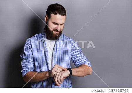 Bearded man clicking on screen of his smart watch. Pleased smiling man enjoying his modern hand clock, gray studio background, copy space 137218705
