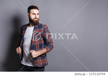 Handsome bearded man looking away, copy space. Portrait of young confident hipster holding his checkered jacket, gray studio background 137218744