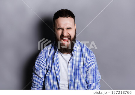 Portrait of angry crying bearded man. Aggressive guy with closed eyes screaming loudly, gray studio background. Psychotherapy exercise Portrait of angry crying bearded man. Aggressive guy with closed eyes screaming loudly, gray studio background. Psychotherapy exercise 137218766