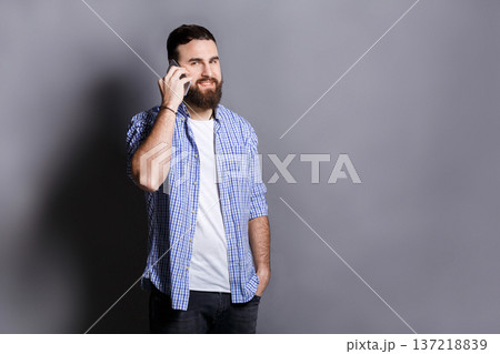 Cheerful bearded man talking on phone. Young smiling guy having nice conversation, gray studio background, copy space. Communication concept 137218839