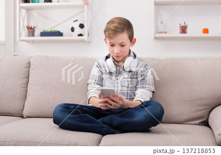 Teenager messaging to friends chat on mobile sitting on couch in living room at home. Stylish kid in checkered shirt and jeans with headphones on the neck 137218853