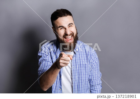 Furious bearded man shouting loud and point at camera. Young angry guy screaming at you, gray studio background 137218901