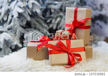 Christmas gifts decorated with red ribbon bows on soft fluffy carpet at blurred snowy fir tree background, closeup, selective focus. Winter holidays concept Christmas gifts decorated with red ribbon bows on soft fluffy carpet at blurred snowy fir tree background, closeup, selective focus. Winter holidays concept 137218981