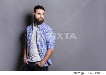 Handsome bearded man posing with hands in pocket. Portrait of young confident guy looking away, gray studio background, copy space Handsome bearded man posing with hands in pocket. Portrait of young confident guy looking away, gray studio background, copy space 137219039
