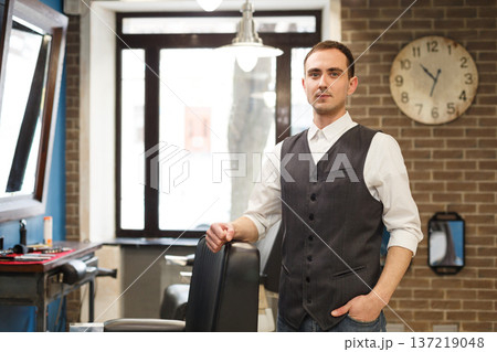Young barber waits for client, looking at camera. Male hairstylist in barbershop Young barber waits for client, looking at camera. Male hairstylist in barbershop 137219048