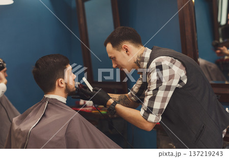 Barber styling beard with trimmer at barbershop. Stylish hairdresser in male hair salon Barber styling beard with trimmer at barbershop. Stylish hairdresser in male hair salon 137219243