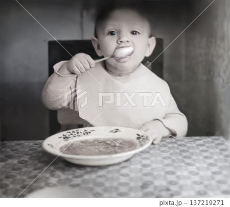A vintage portrait of a little boy eating from a plate with a spoon. Retro photo from the 1960s. 137219271