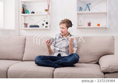 Excited teenage boy with braces playing football game on smartphone and listening to music in headphones, enjoying win while sitting on sofa in living room at home 137219364