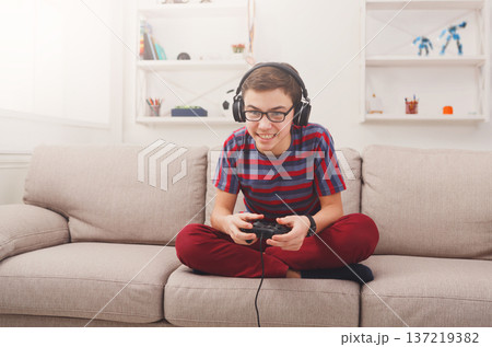 Online gaming concept. Teenage boy playing football video game with joystick wearing headphones while sitting on sofa in living room at home Online gaming concept. Teenage boy playing football video game with joystick wearing headphones while sitting on sofa in living room at home 137219382
